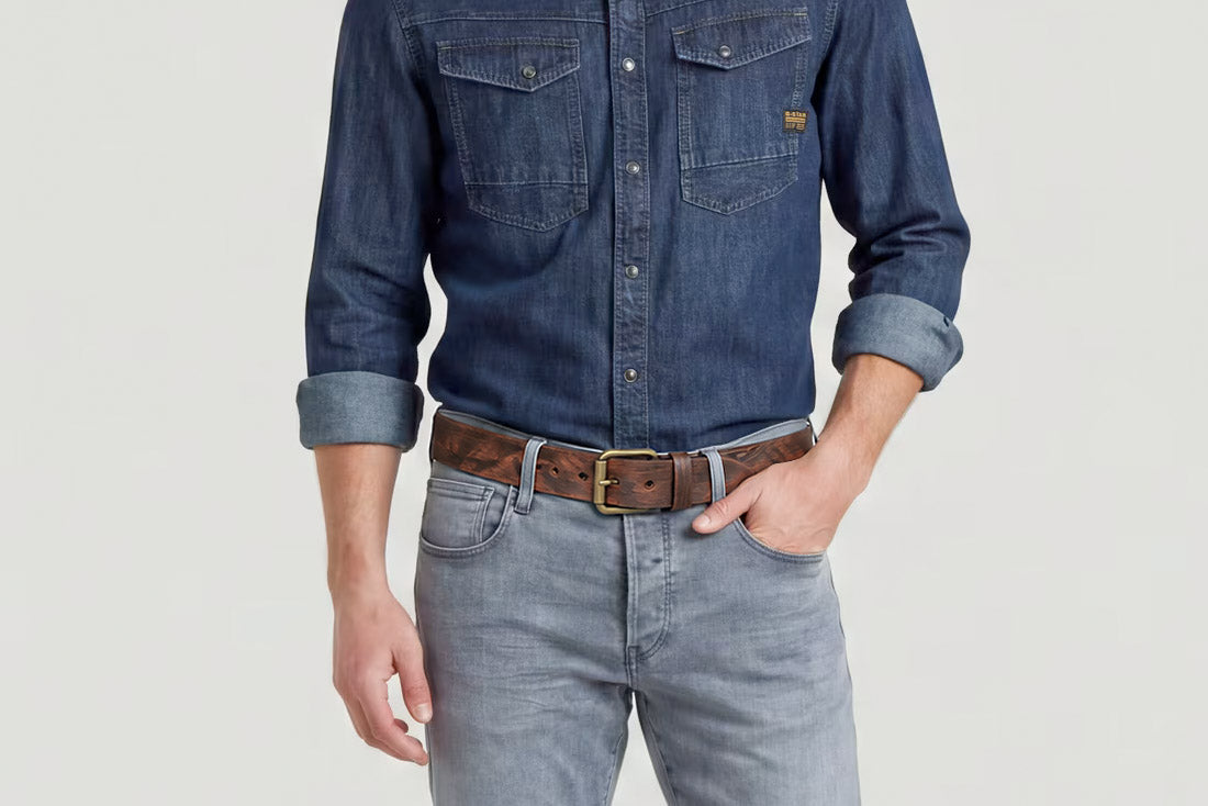 Men’s brown distressed leather belt with antique gold-tone buckle styled with a dark denim shirt and grey jeans.