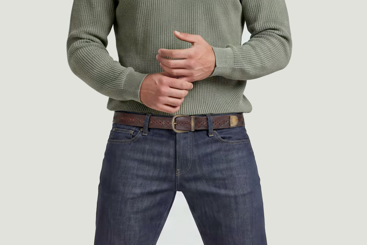 Men's brown perforated leather belt with antique gold buckle and metal keeper, styled with dark jeans and a sage green knit sweater.